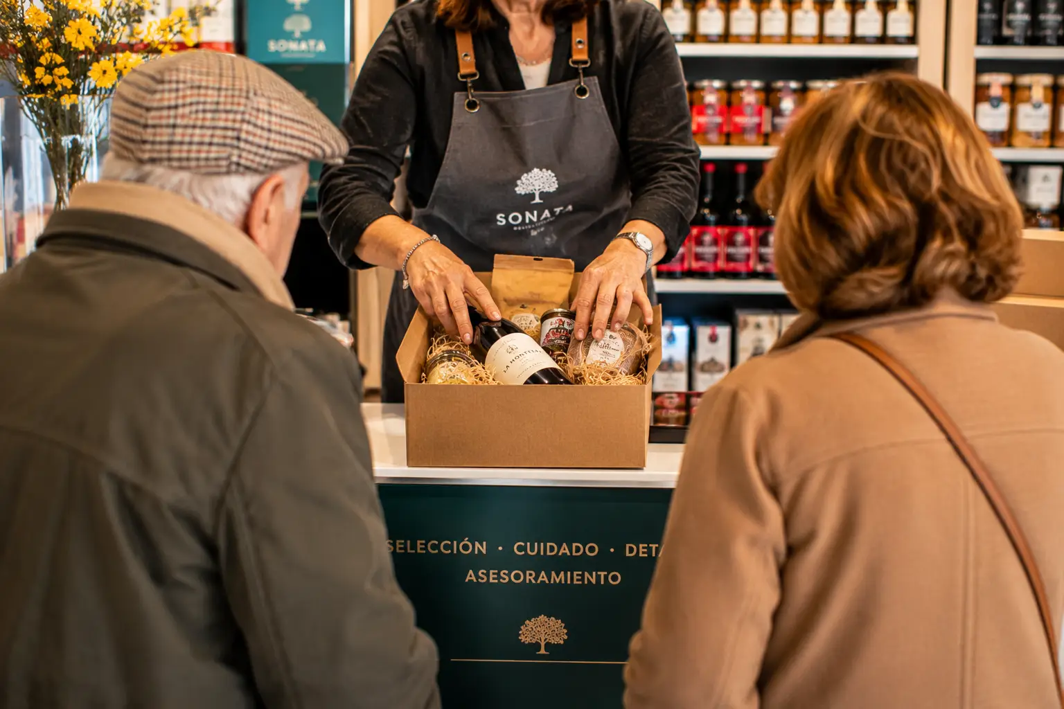 Dos clientes atendidos en el mostrador mientras se prepara una cesta con vino y conservas